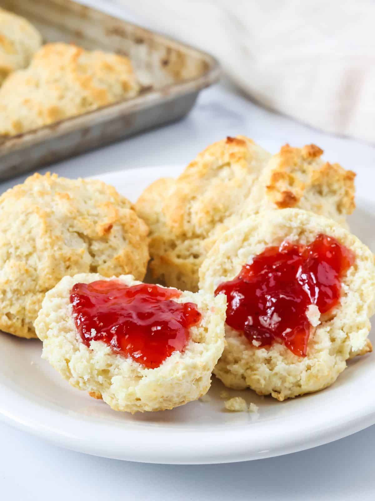 Bisquick drop biscuits served on a white plate. One biscuit is split open and topped with strawberry jam, showing the fluffy interior of the biscuit.