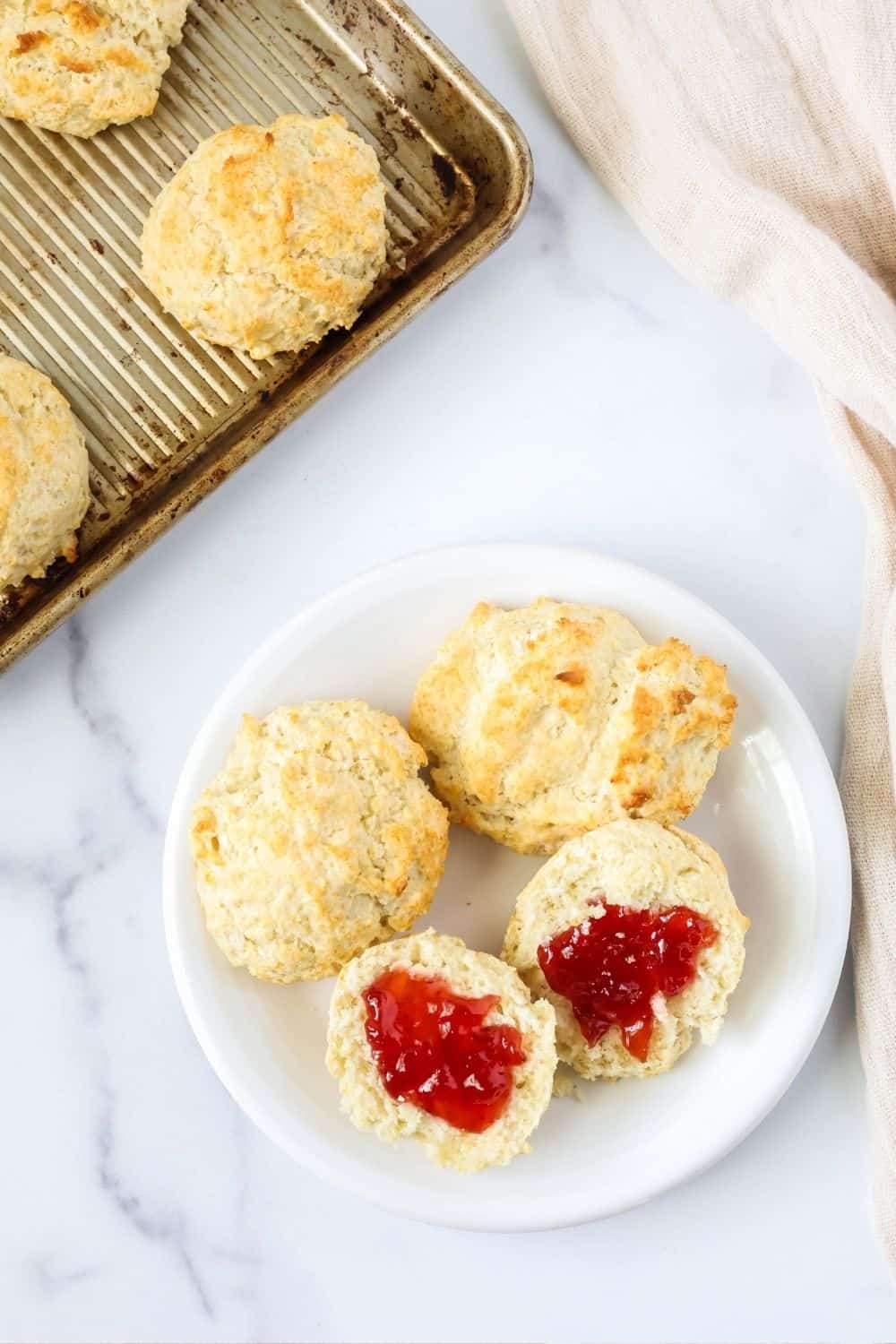 overhead view of a white plate with easy drop biscuits made with Bisquick on the plate. One biscuit is split in half and spread with jam. The pan of remaining biscuits is in the background.