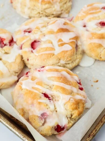 Several fresh strawberry biscuits, made with Bisquick and topped with glaze, on a parchment-lined baking sheet.