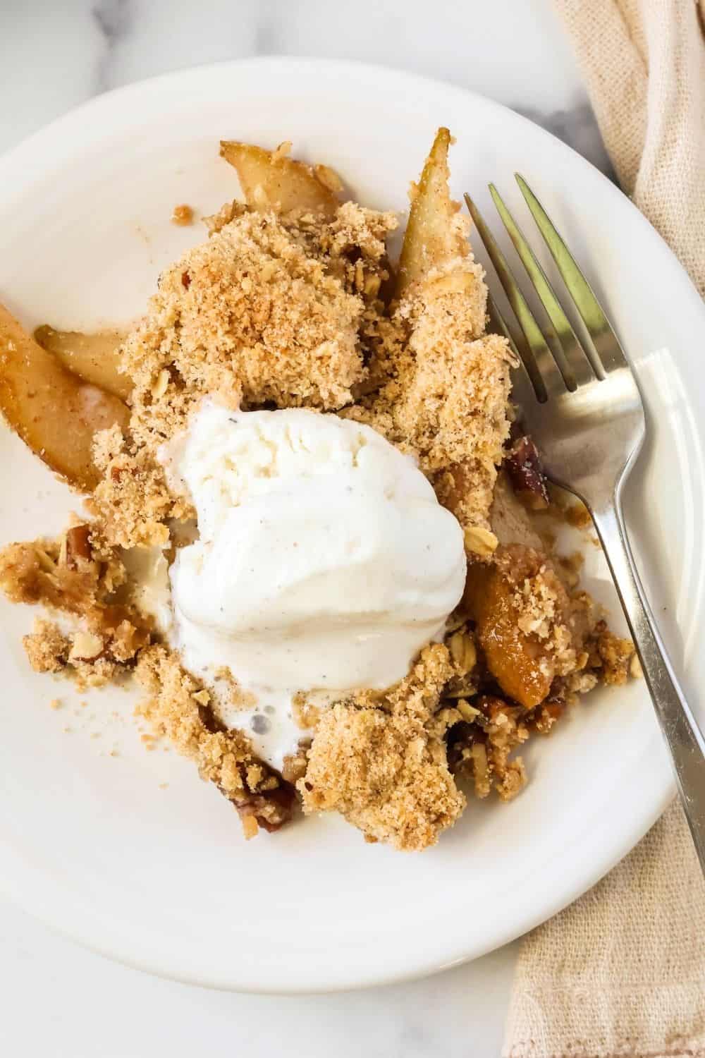 overhead view of cinnamon pear crumble served on a white plate, topped with ice cream. A fork is on the plate.