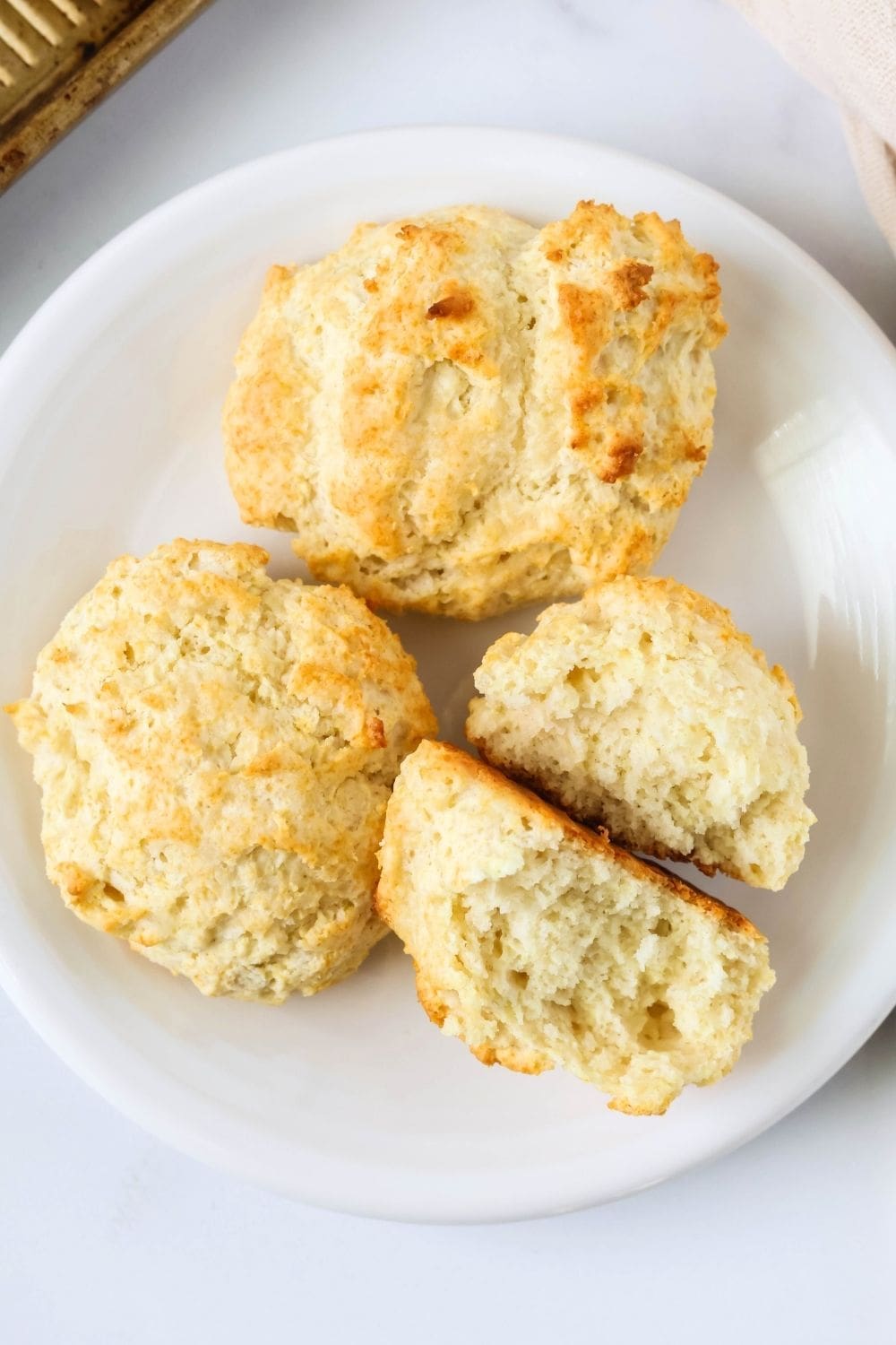 overhead view of three drop biscuits on a white plate. One biscuit is torn in half, showcasing the fluffy interior texture.