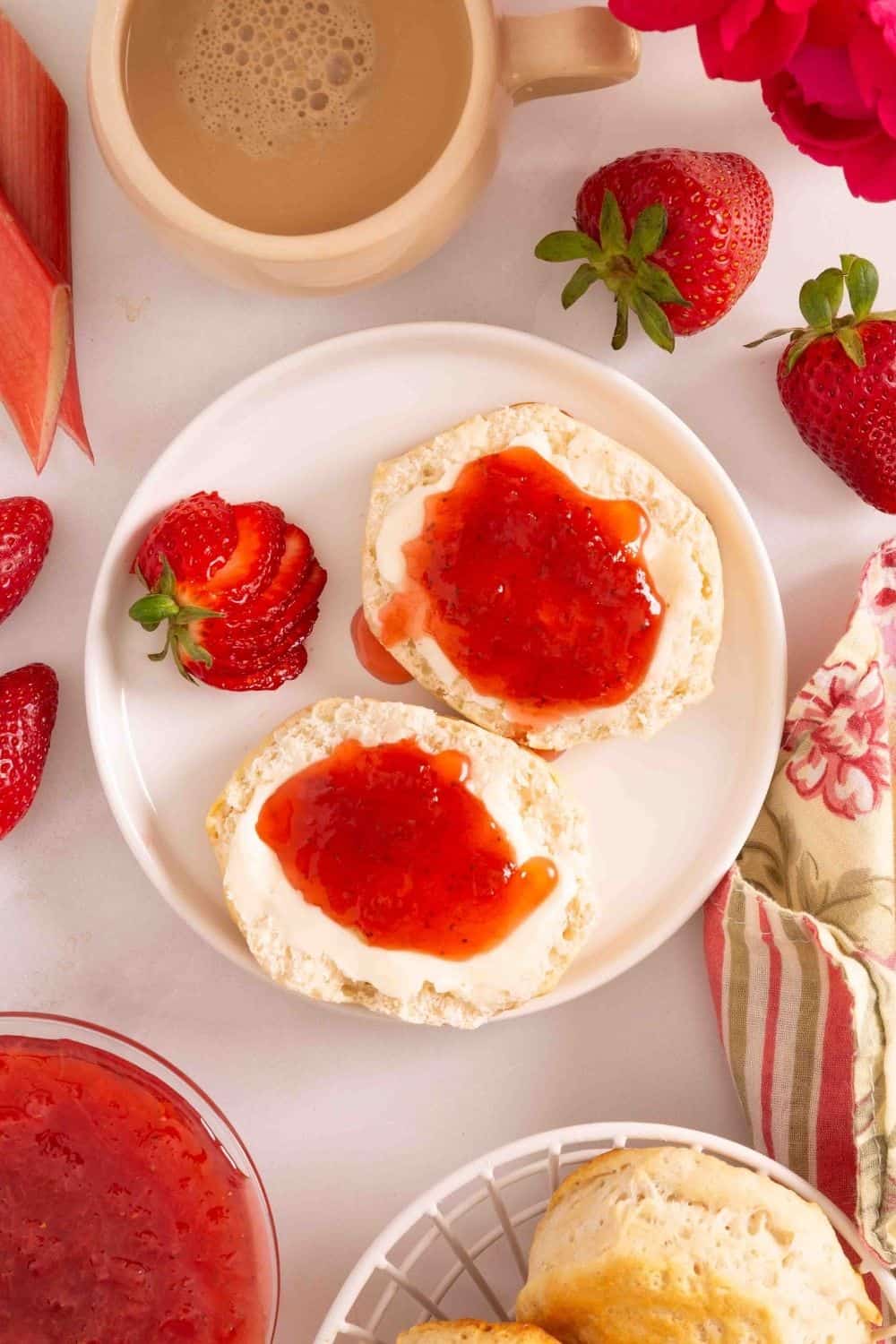 Overhead view of two biscuit halves on a white plate; the biscuits are topped with homemade strawberry rhubarb jam. Fresh strawberries are around the plate, as well as more biscuits and a cup of coffee.