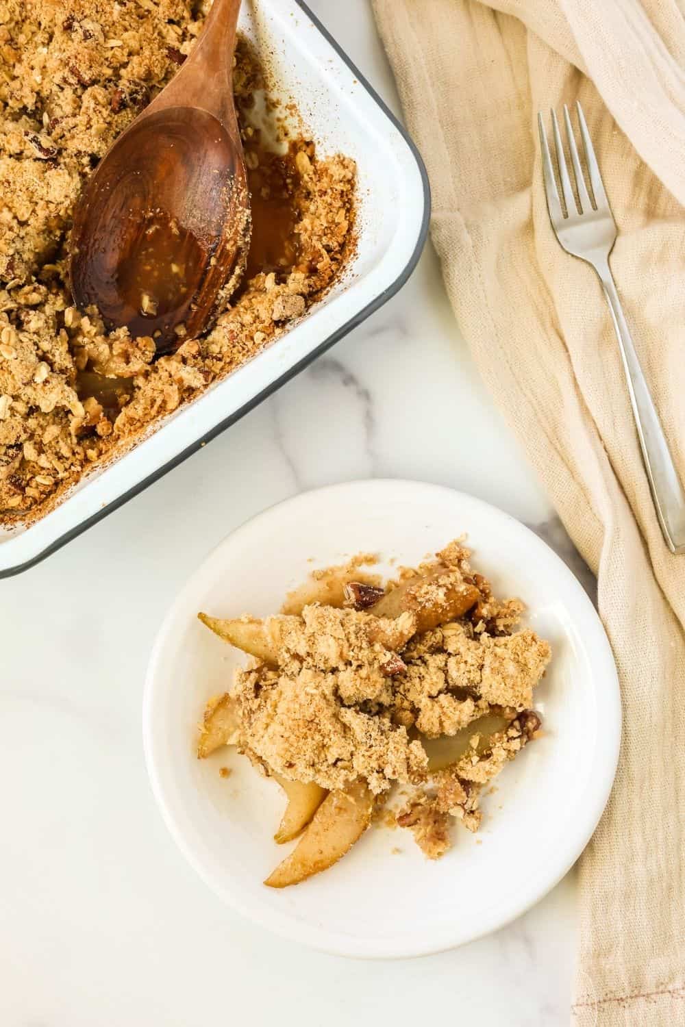 overhead view of a small dessert plate with a serving of pear cobbler with crumble topping. The pan of pear crisp is in the background.