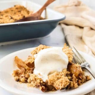 Pear crisp served with a scoop of vanilla ice cream, on a white plate with a fork. The pan of remaining pear crisp is in the background.