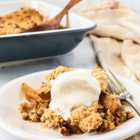 Pear crisp served with a scoop of vanilla ice cream, on a white plate with a fork. The pan of remaining pear crisp is in the background.