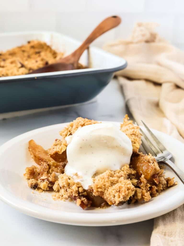 Pear crisp served with a scoop of vanilla ice cream, on a white plate with a fork. The pan of remaining pear crisp is in the background.