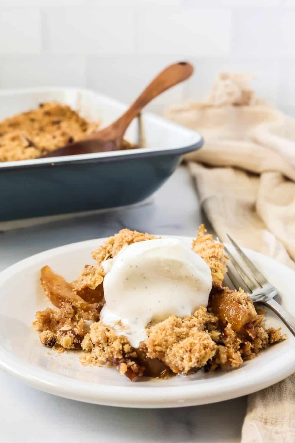 A serving of pear crisp made with fresh pears on a white plate, topped with vanilla ice cream. The pan of remaining crisp is in the background.