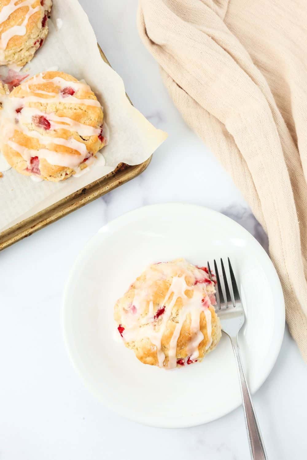 overhead view of a strawberry biscuit on a white plate with a fork. A pan of fresh strawberry biscuits is in the background.