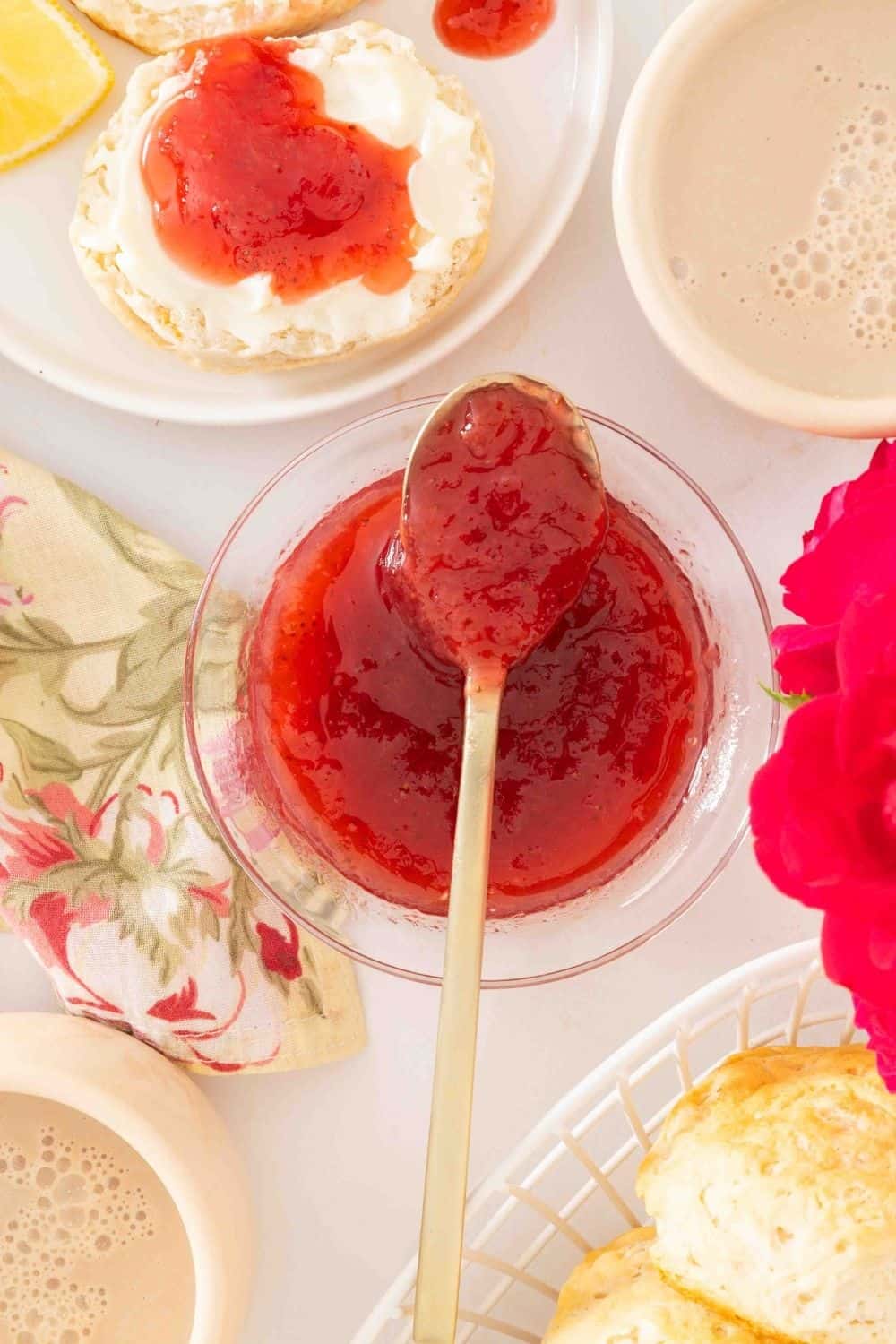Overhead view of a glass dish of strawberry and rhubarb jam. A spoon is lifting out some of the jam from the dish. A biscuit topped with jam is in the background.
