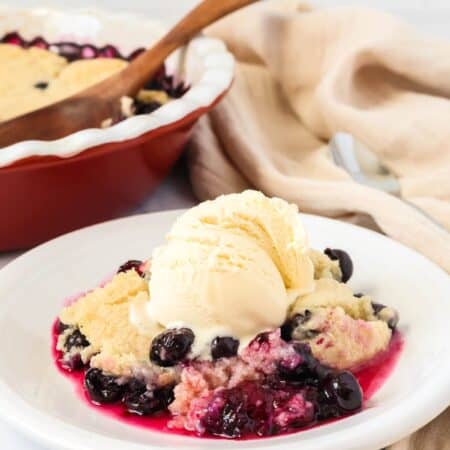 Bisquick blueberry cobbler served on a white plate with a scoop of vanilla ice cream. The baking dish is in the background.