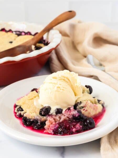 Bisquick blueberry cobbler served on a white plate with a scoop of vanilla ice cream. The baking dish is in the background.