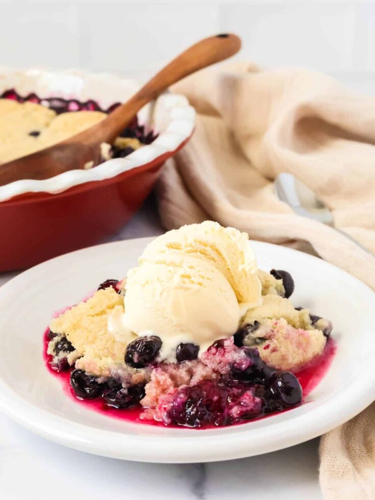 Bisquick blueberry cobbler served on a white plate with a scoop of vanilla ice cream. The baking dish is in the background.