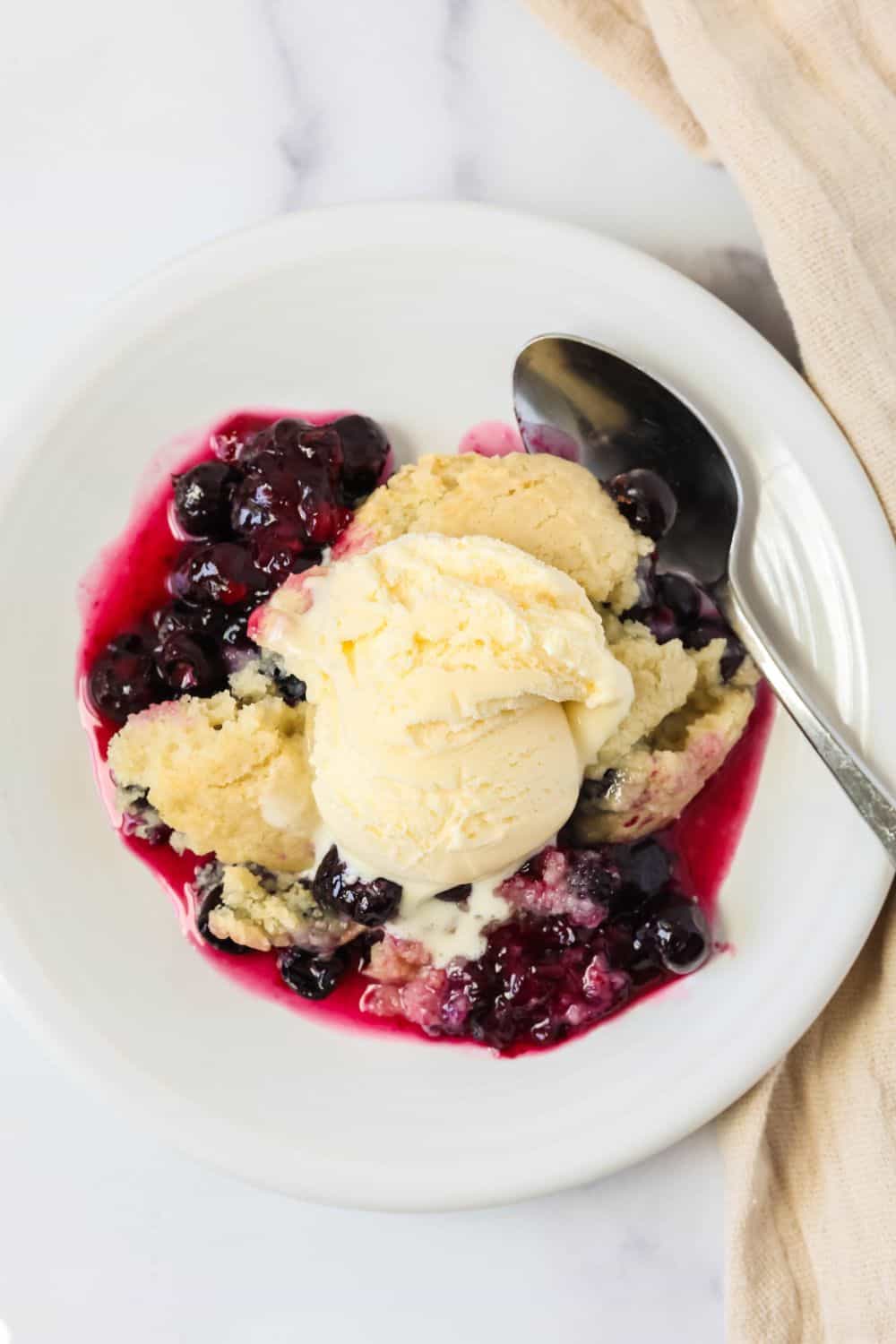 Overhead view of a plate of Bisquick fresh blueberry cobbler topped with vanilla ice cream. A spoon is next to the cobbler.