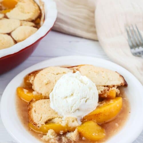 Bisquick peach cobbler on a white plate, topped with ice cream. The baking dish of cobbler is in the background.