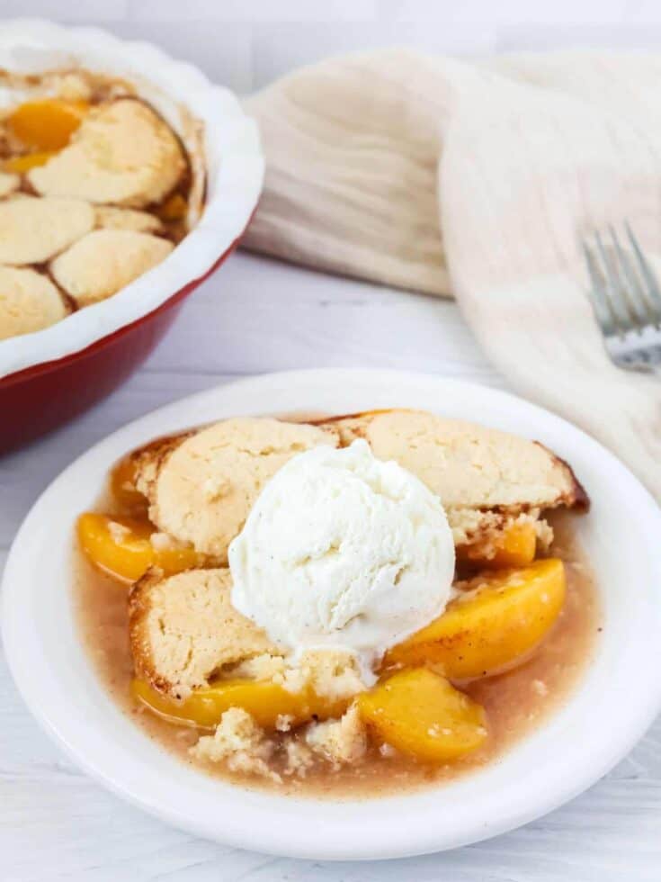 Bisquick peach cobbler on a white plate, topped with ice cream. The baking dish of cobbler is in the background.