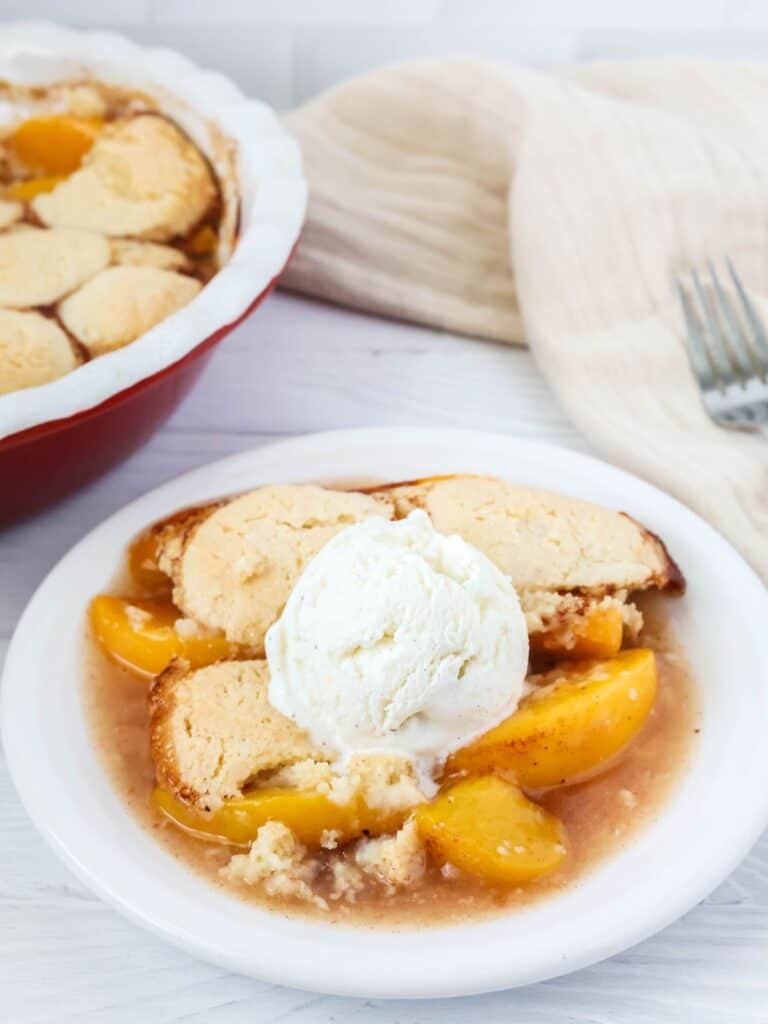 Bisquick peach cobbler on a white plate, topped with ice cream. The baking dish of cobbler is in the background.