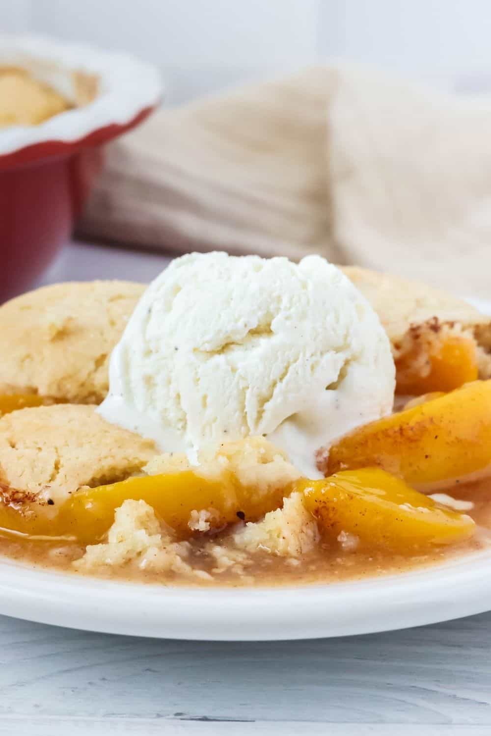 side view of a helping of canned peach cobbler made with Bisquick, served on a white plate, topped with a scoop of vanilla ice cream that's beginning to melt from the warm cobbler.