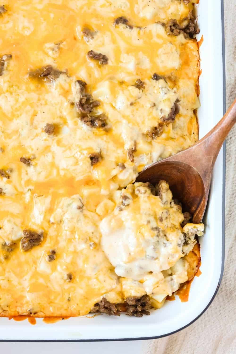 Baking dish of a casserole of cheesy ground beef and hash brown potatoes. A wooden spoon is scooping out a serving.