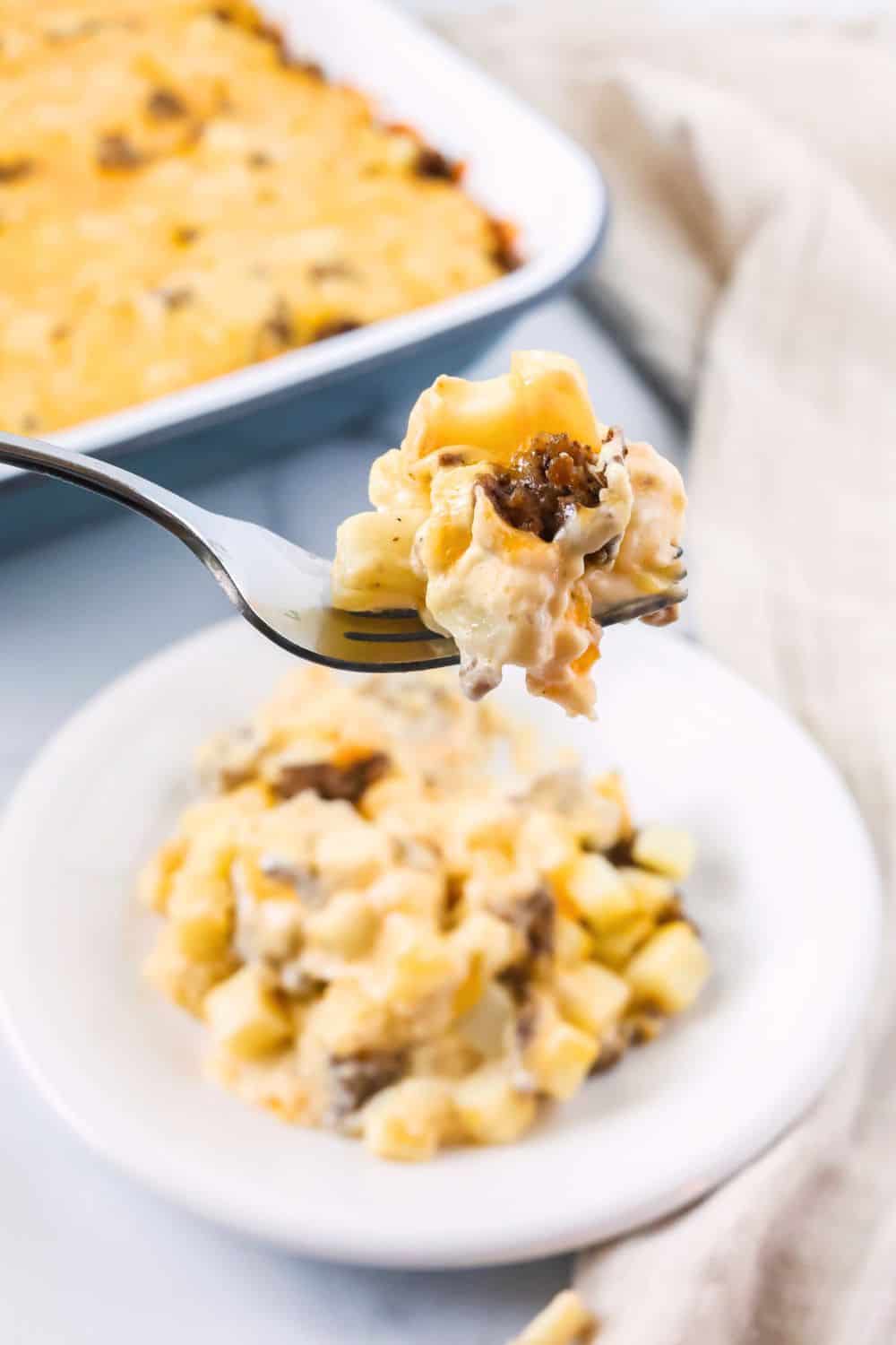 A fork lifts a bite of cheesy hobo casserole with ground beef from a plate. The casserole dish is in the background.