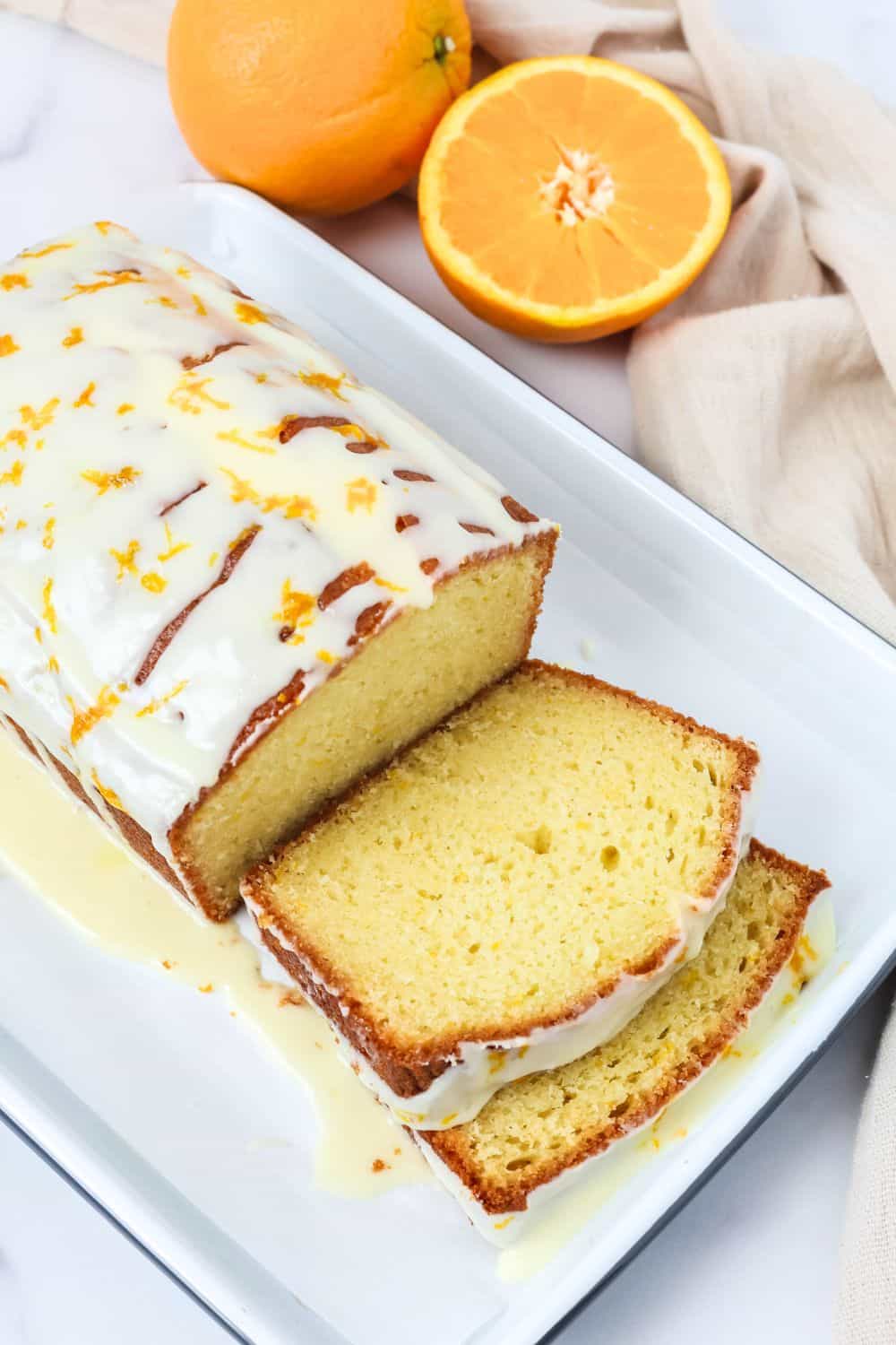 overhead view of a partially sliced easy orange loaf, topped with glaze, on a white rectangular platter. Two oranges are next to the platter.