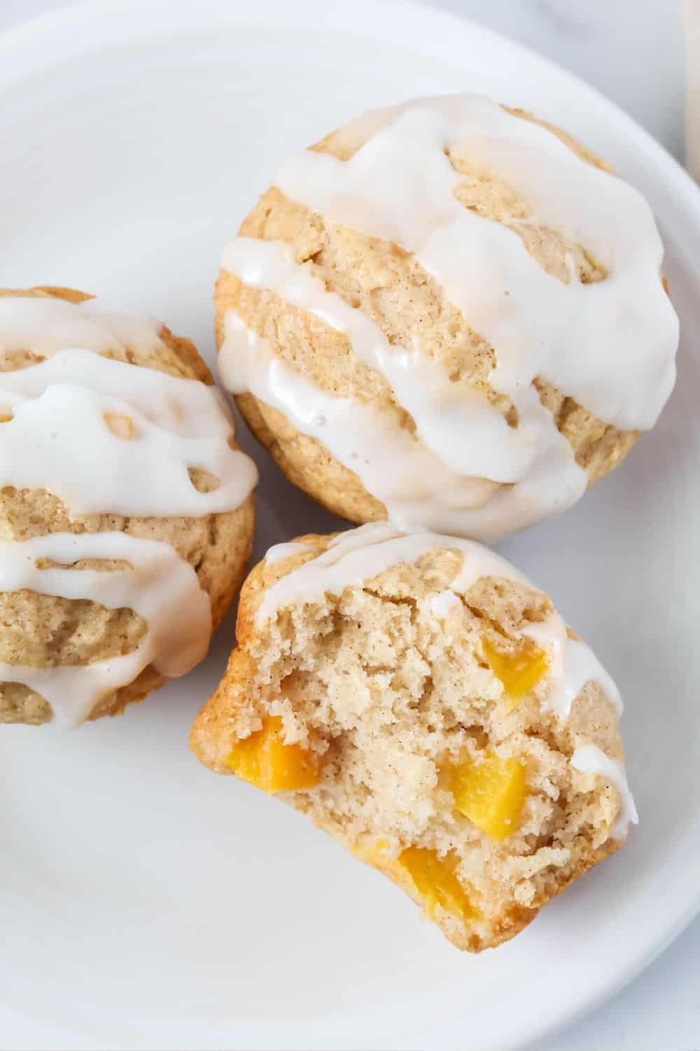 three easy peach muffins on a white plate, topped with icing. One muffin has a bite taken out of it, showing chunks of yellow peaches in the fluffy interior.