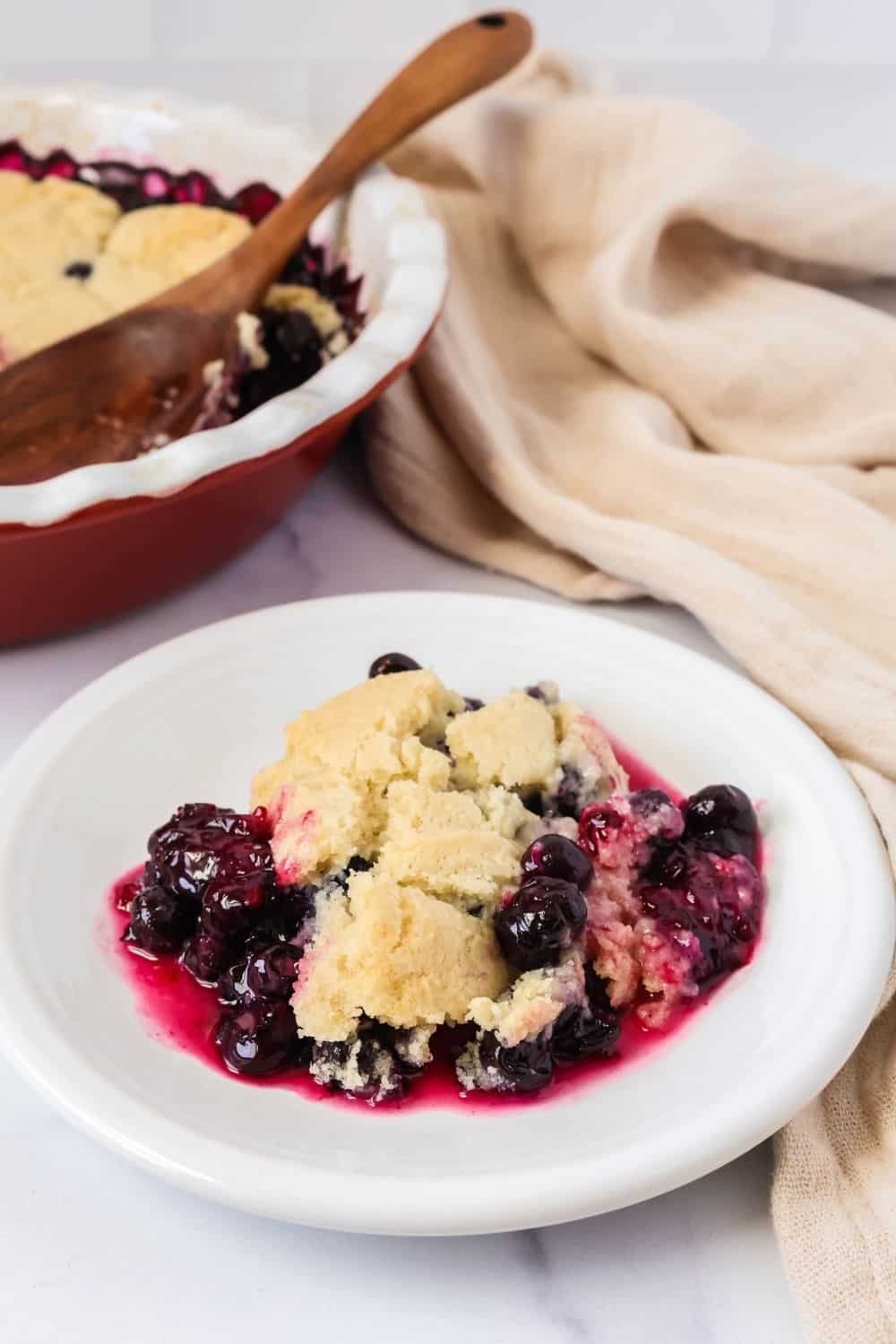 A serving of fresh blueberry cobbler with biscuit-like topping on a white plate, with the dish of remaining cobbler in the background.
