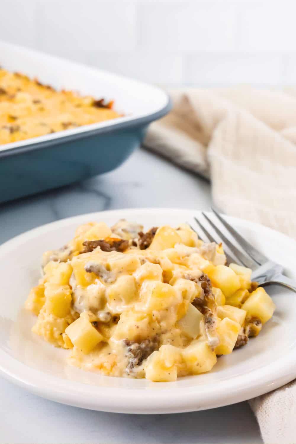 A white plate with a helping of ground beef hobo casserole on it, with a fork on the plate. The casserole dish is in the background.