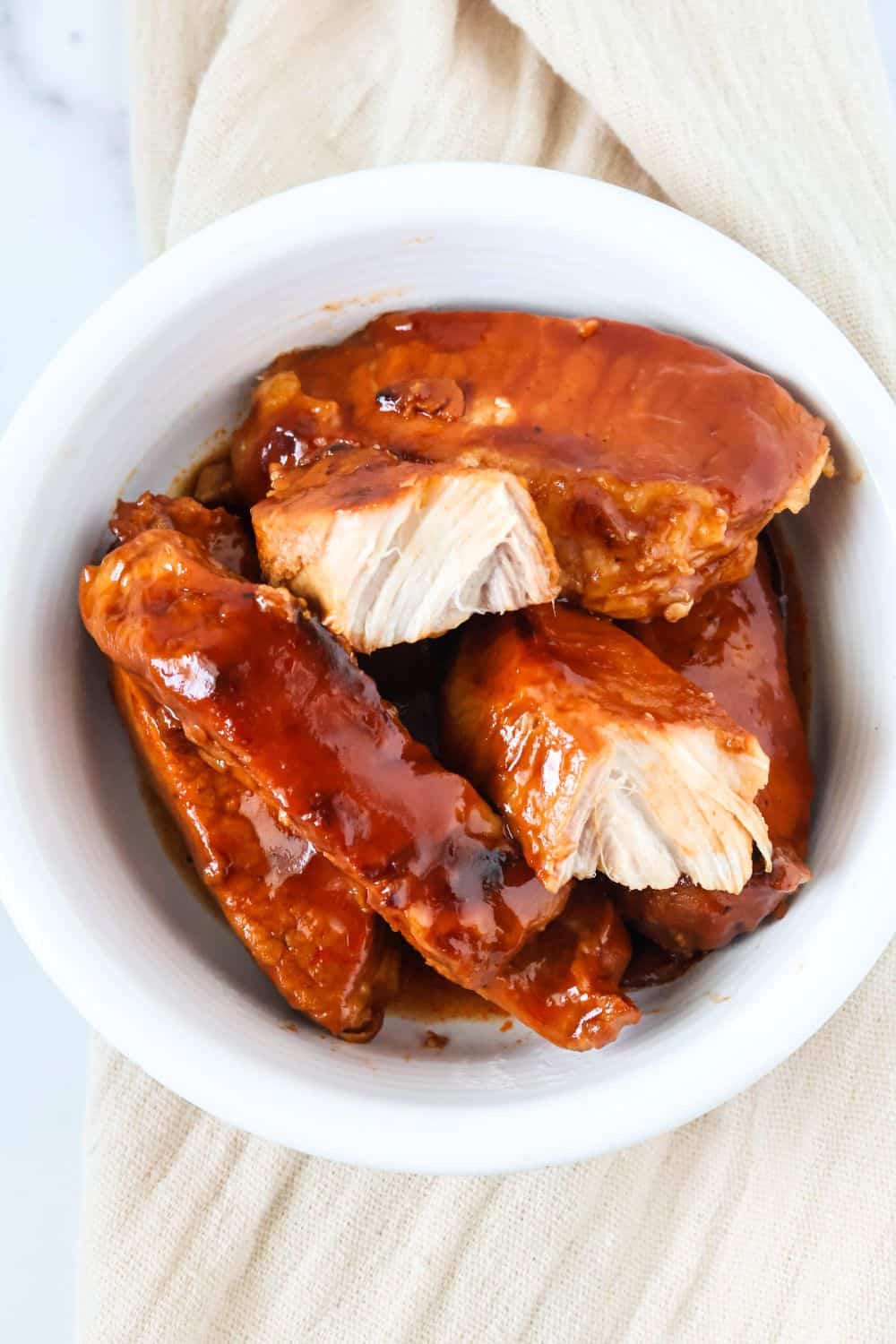 Overhead view of a white bowl filled with Instant Pot boneless ribs, smothered in BBQ sauce. One of the pork ribs has been torn apart, showcasing the moist meat inside.