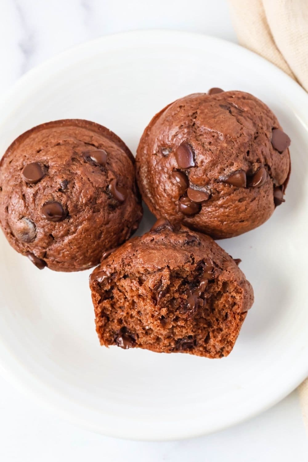 overhead view of three moist chocolate muffins made using Bisquick. One muffin has a bite taken out of it, showing the tender interior and melty chocolate chips.