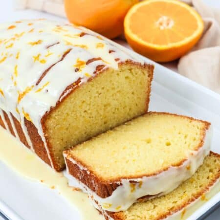 Orange bread loaf cake, partially sliced and topped with glaze, on a white platter. Two fresh oranges are in the background.