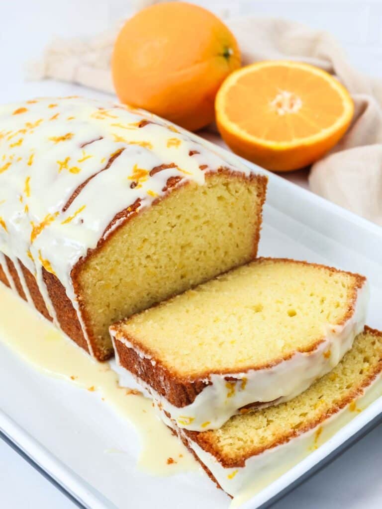 Orange bread loaf cake, partially sliced and topped with glaze, on a white platter. Two fresh oranges are in the background.
