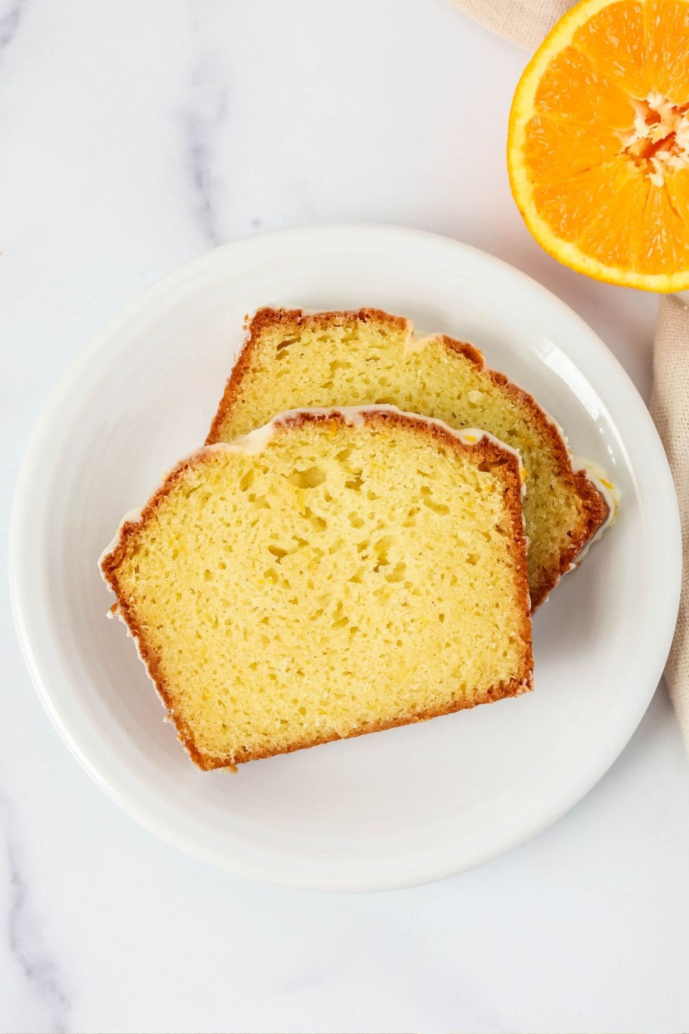 Two slices of orange loaf cake on a white plate. An orange is in the background.