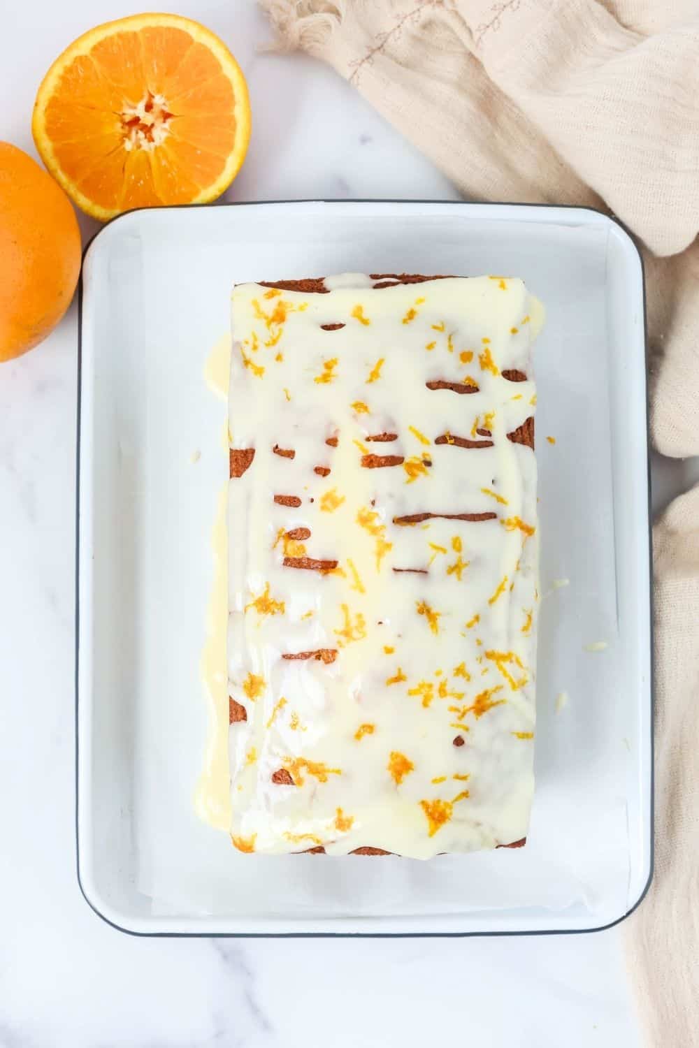 overhead view of orange loaf cake topped with icing, on a rectangular plate with two oranges next to it.