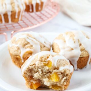 Three peach muffins made with Bisquick served on a white plate, with more muffins on a wire rack in the background.