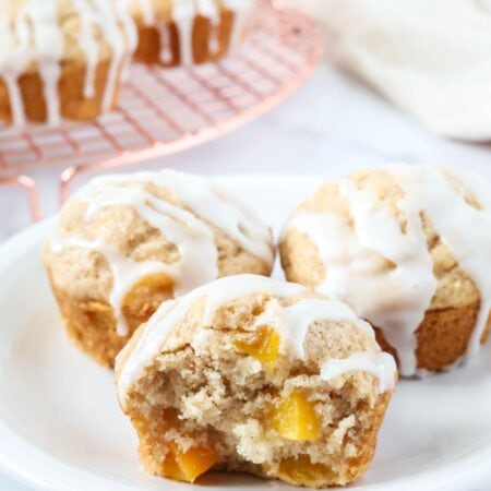 Three peach muffins made with Bisquick served on a white plate, with more muffins on a wire rack in the background.