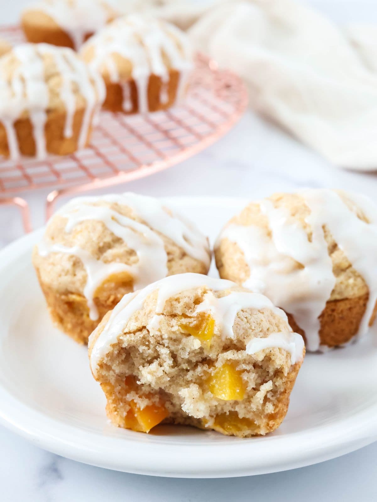 Three peach muffins made with Bisquick served on a white plate, with more muffins on a wire rack in the background.