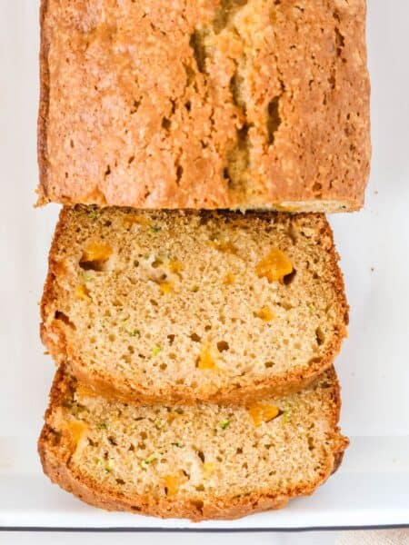 Peach zucchini loaf on a white platter, partially sliced, showing the moist interior of the bread dotted with zucchini pieces and diced peaches.