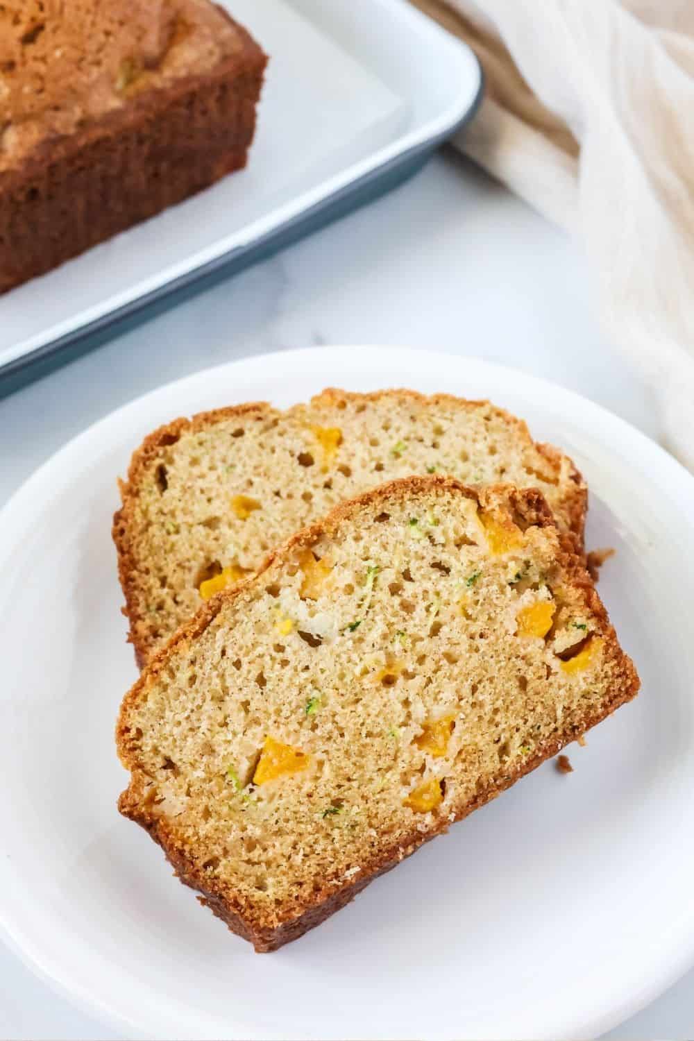 Overhead view of two slices of peach zucchini bread made with canned or fresh peaches, served on a white plate with the remainder of the loaf of quickbread in the background.