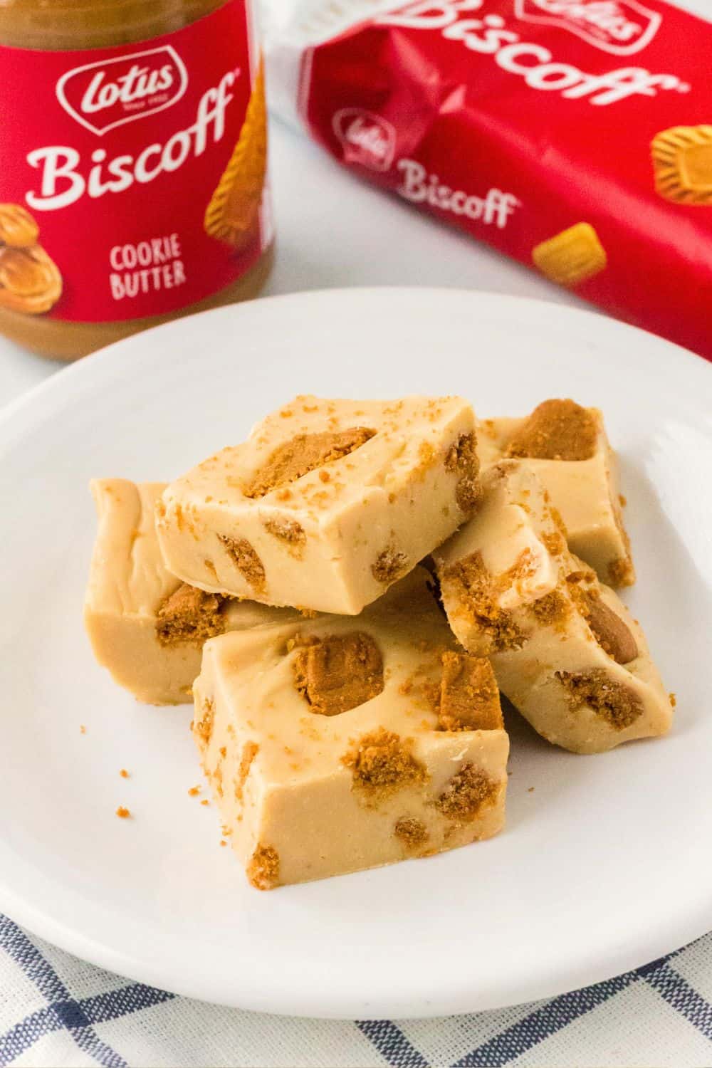 Several pieces of Biscoff fudge on a white plate. A jar of Biscoff cookie butter and a package of Biscoff cookies are in the background.