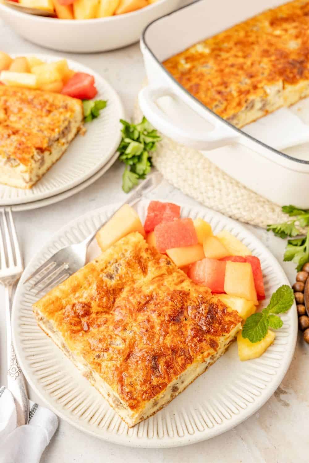 Slice of cheesy sausage egg breakfast casserole on a white plate, served with fresh fruit. The baking dish of casserole is in the background.