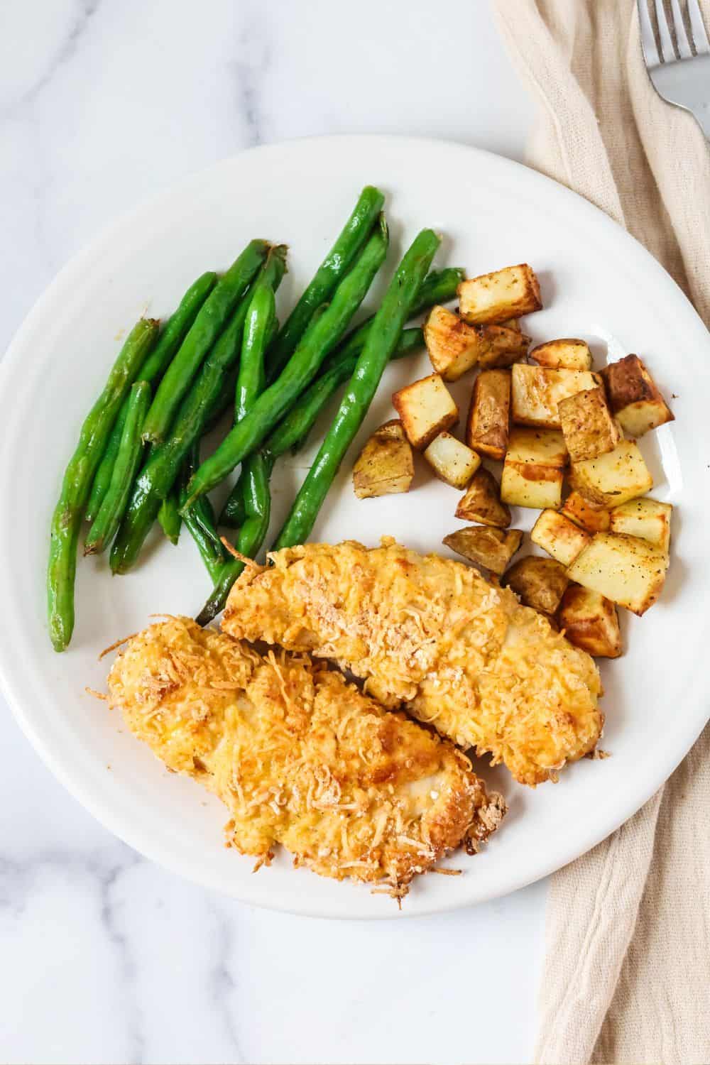 Overhead view of a white plate containing baked chicken tenders breaded with Bisquick, alongside diced potatoes and sauteed green beans.
