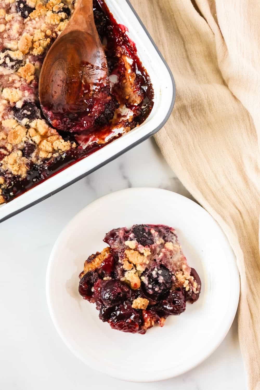 overhead view of a serving of cherry cobbler on a white plate. The baking dish of cobbler and a wooden serving spoon are in the background.