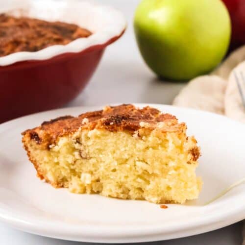 Country apple cake slice served on a white plate, with the cake pan and fresh apples in the background.