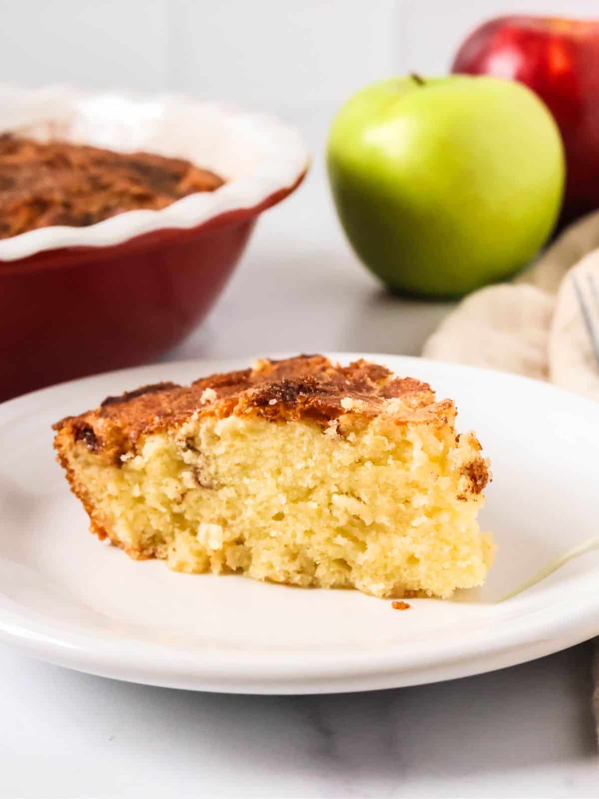 Country apple cake slice served on a white plate, with the cake pan and fresh apples in the background.