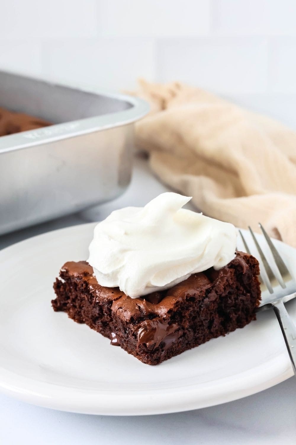 Slice of low fat chocolate dump cake on a white plate. The slice is garnished with Cool Whip. A cake pan is in the background.