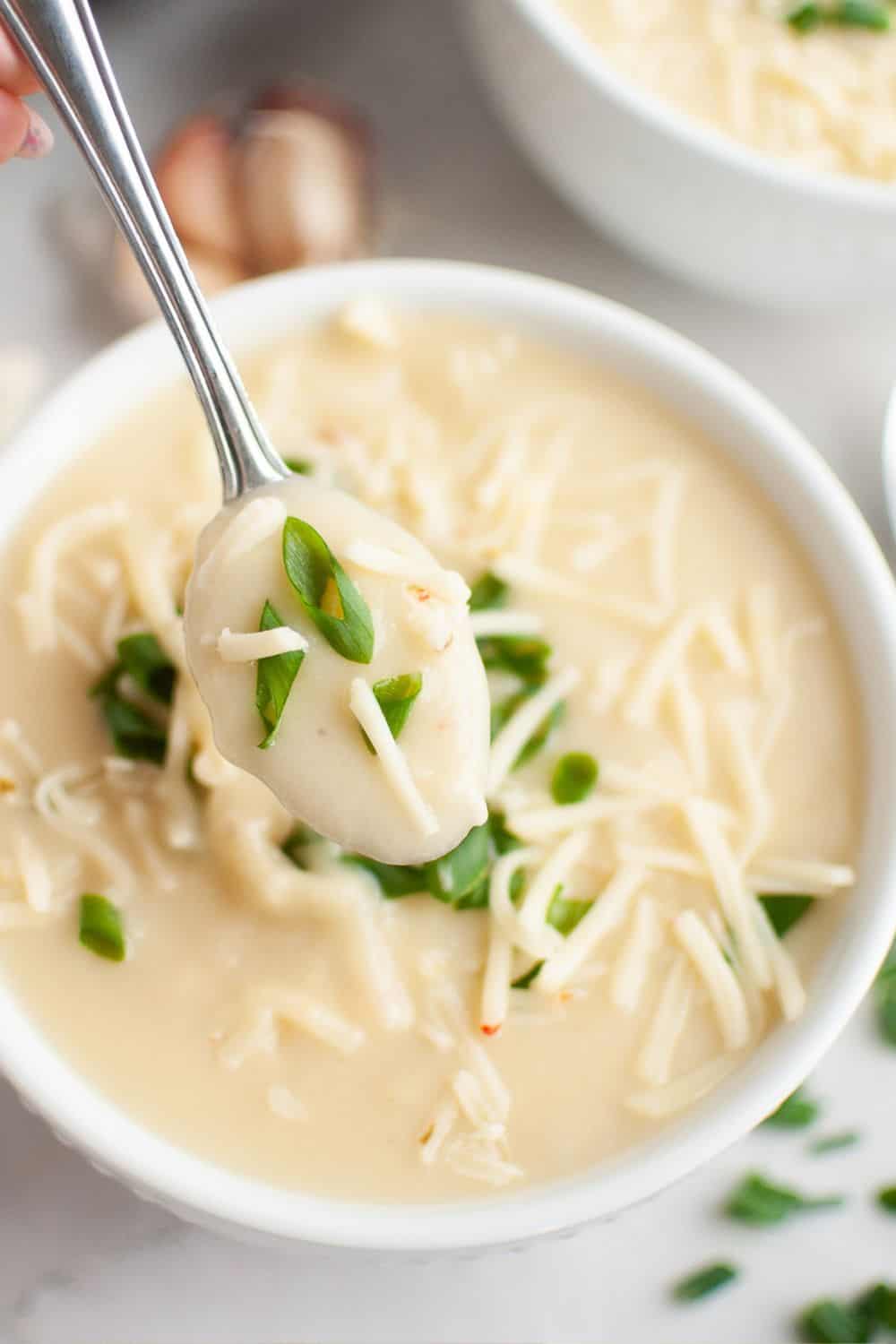 A spoon scoops out a bite of cauliflower soup topped with cheese and green onions.