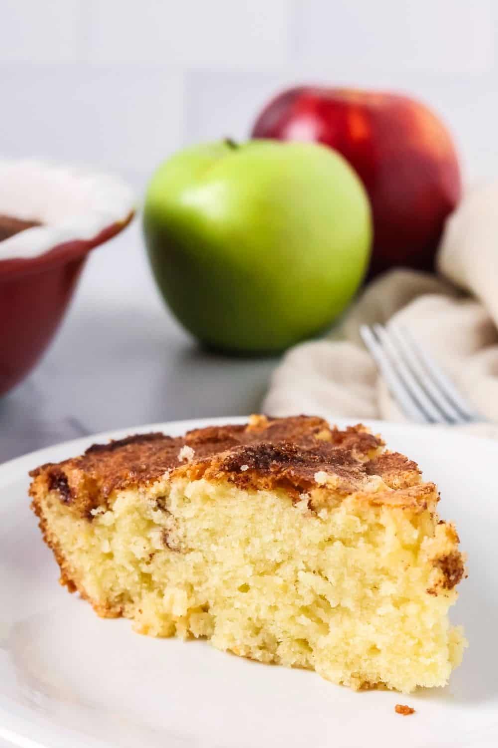 Slice of apple cake made with grandma's recipe served on a white plate. Two apples are in the background.