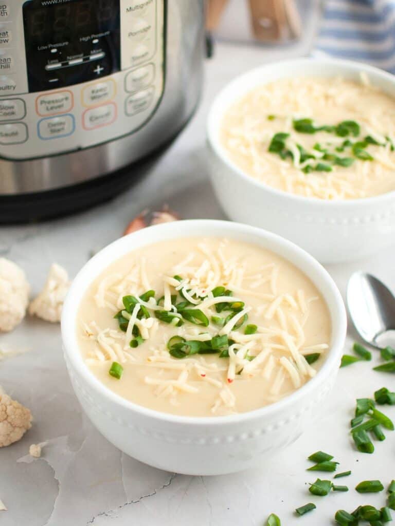 Instant Pot cheesy cauliflower soup in white bowls, with an Instant Pot pressure cooker in the background.