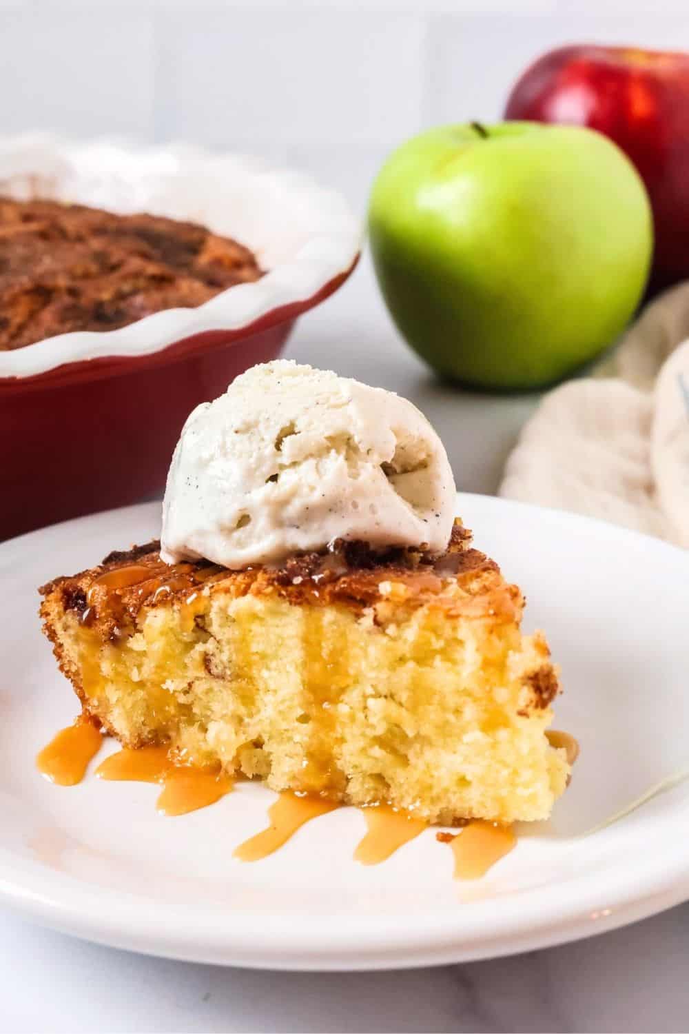 Slice of apple cake made with fresh apples, topped with ice cream and caramel sauce. The cake pan and two fresh apples are in the background.
