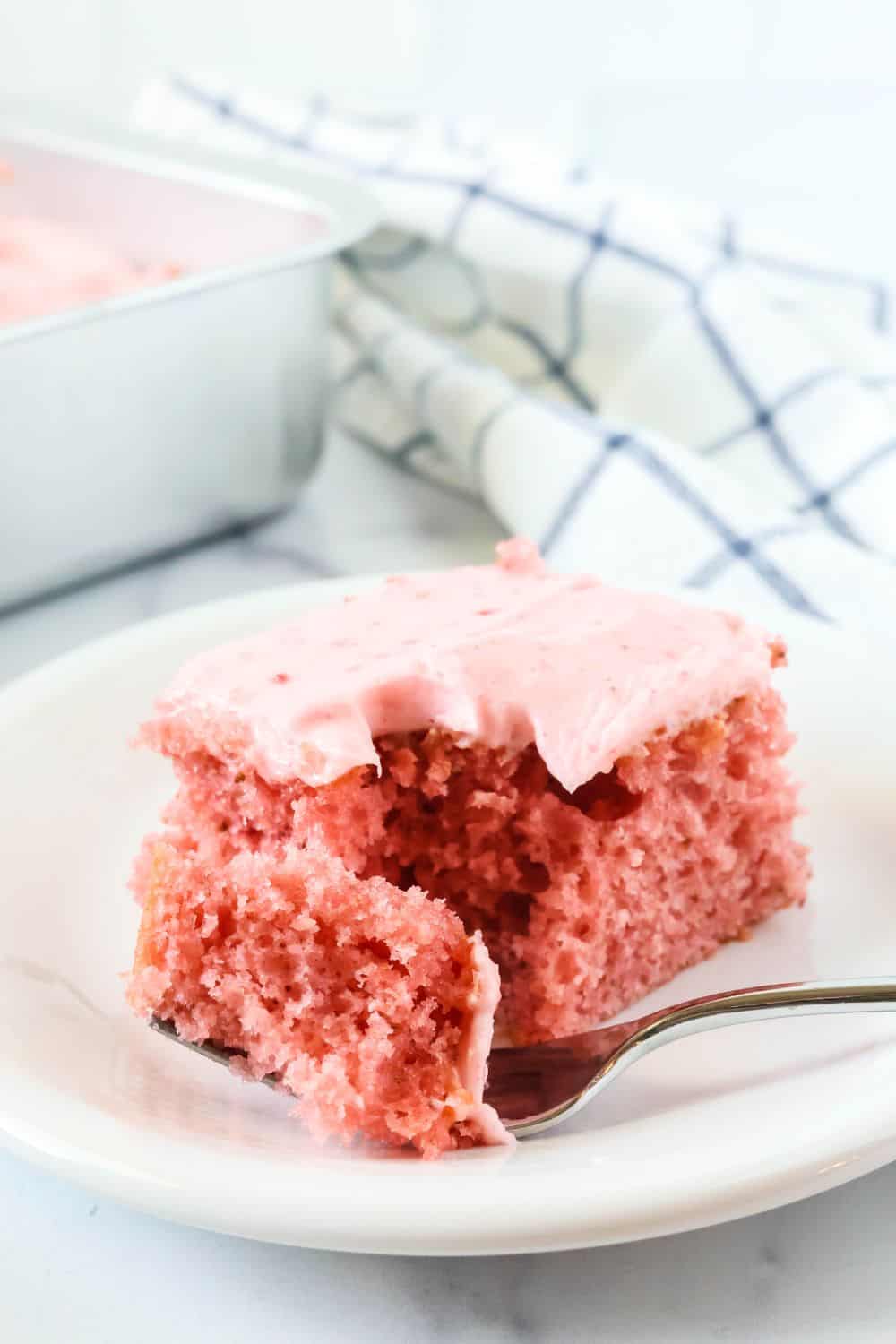 Slice of strawberry cake with strawberry cream cheese frosting on a white plate with a fork. The sheet pan of remaining cake is in the background.